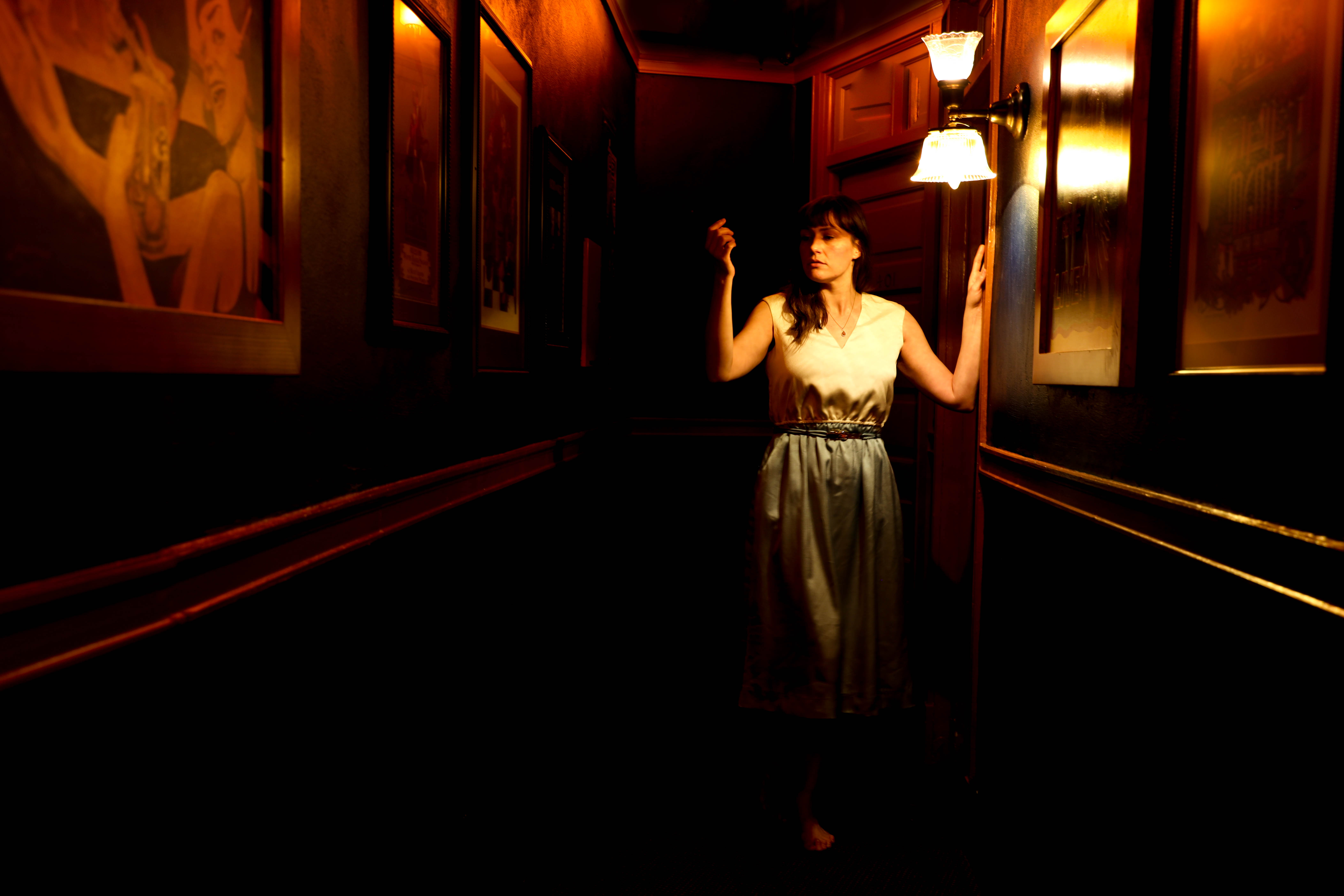 Stephanie Strange standing in a dark hallway with many framed
               pictures on the wall, holding one arm up to look at her hand, wearing
               a white and blue full length dress