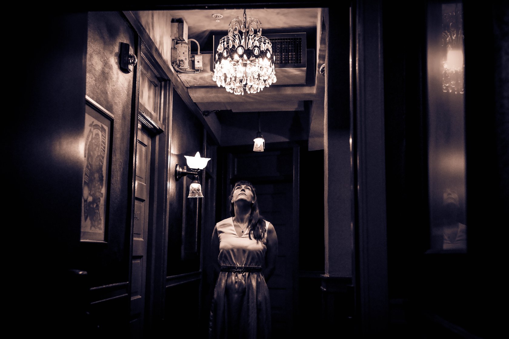 A sepia toned photo of Stephanie Strange looking up towards
               an oranate chandelier in a thin hallway with antique lamp
               fixtures and framed photos