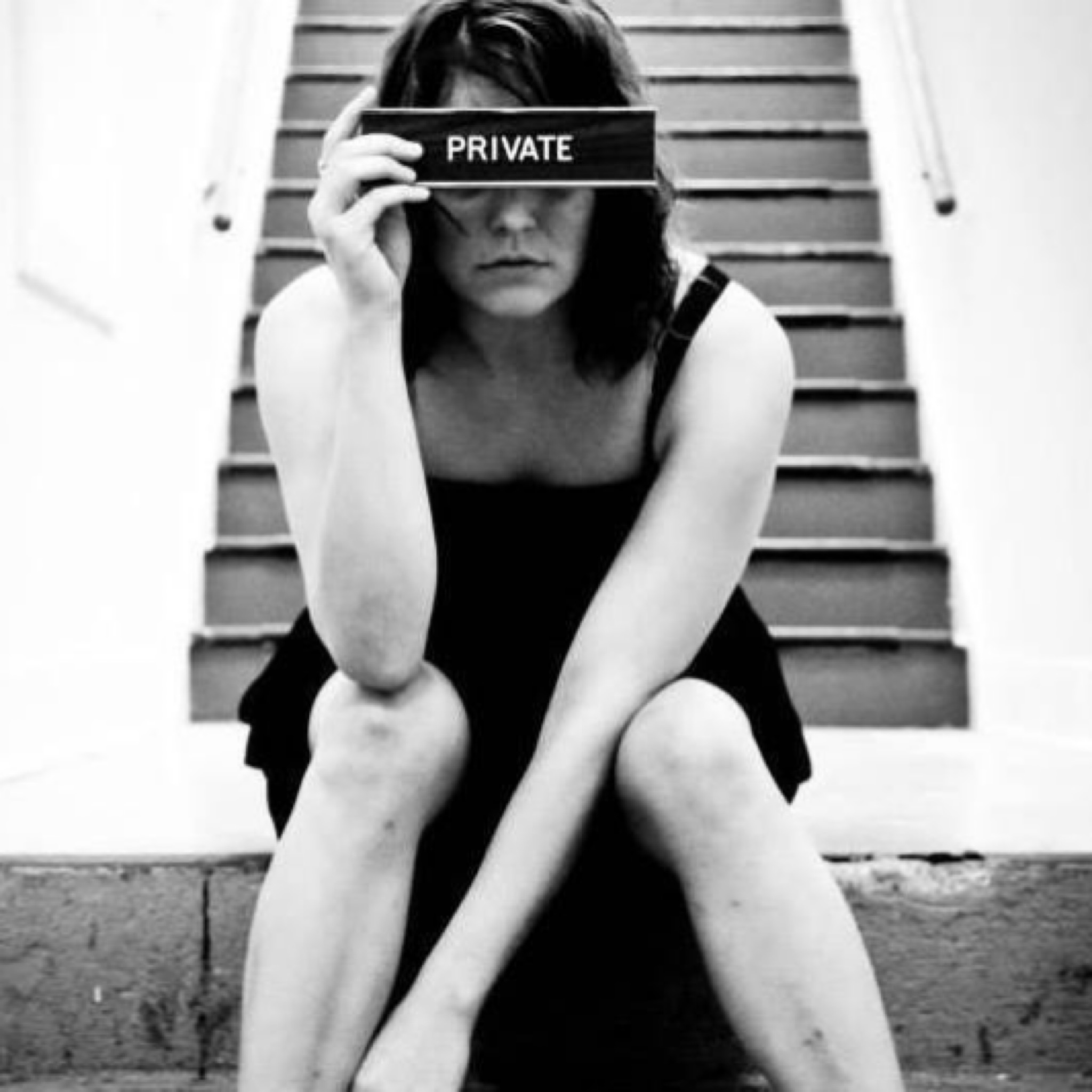 Black and white photo of Stephanie Strange sitting on concrete steps holding a sign that says private over her eyes